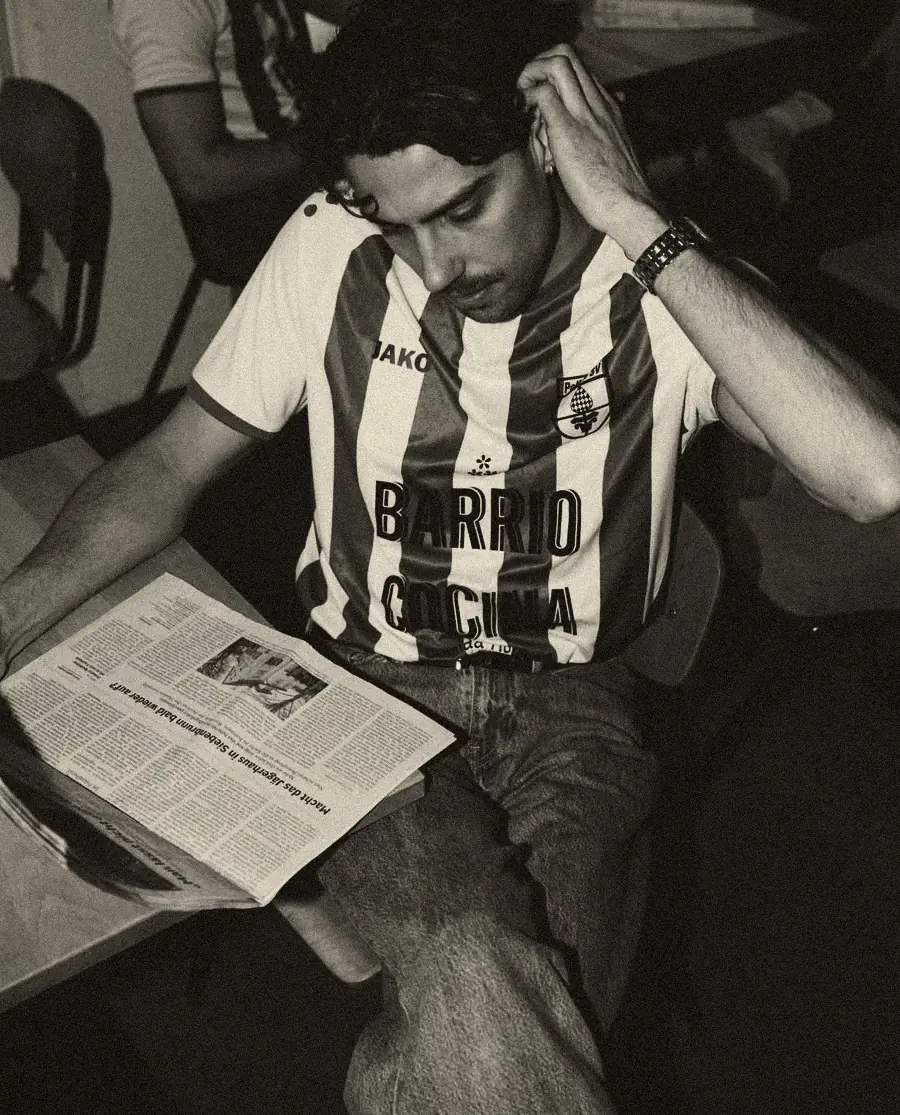 Ein Mann im gestreiften Trikot mit dem Schriftzug Barrio Cocina sitzt am Tisch und liest Zeitung, die Hand im Haar.