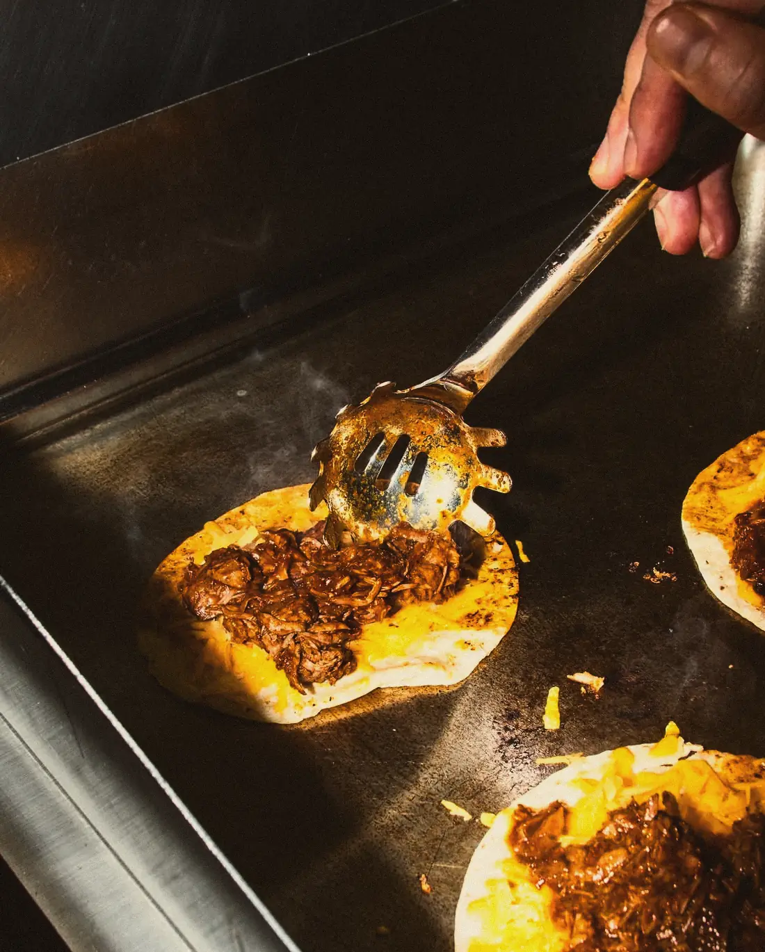 Auf der heißen Grillplatte liegen Tortillas mit Käse und geschmortem Rindfleisch, eine Hand verteilt das Fleisch mit der Zange.