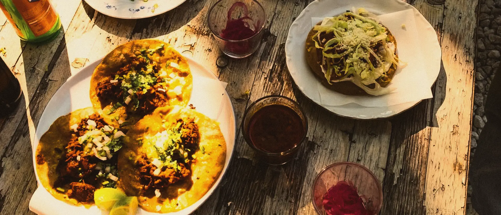 Ein alter Holztisch im Abendlicht voller Tacos, Salsas und eingelegter roter Zwiebeln.